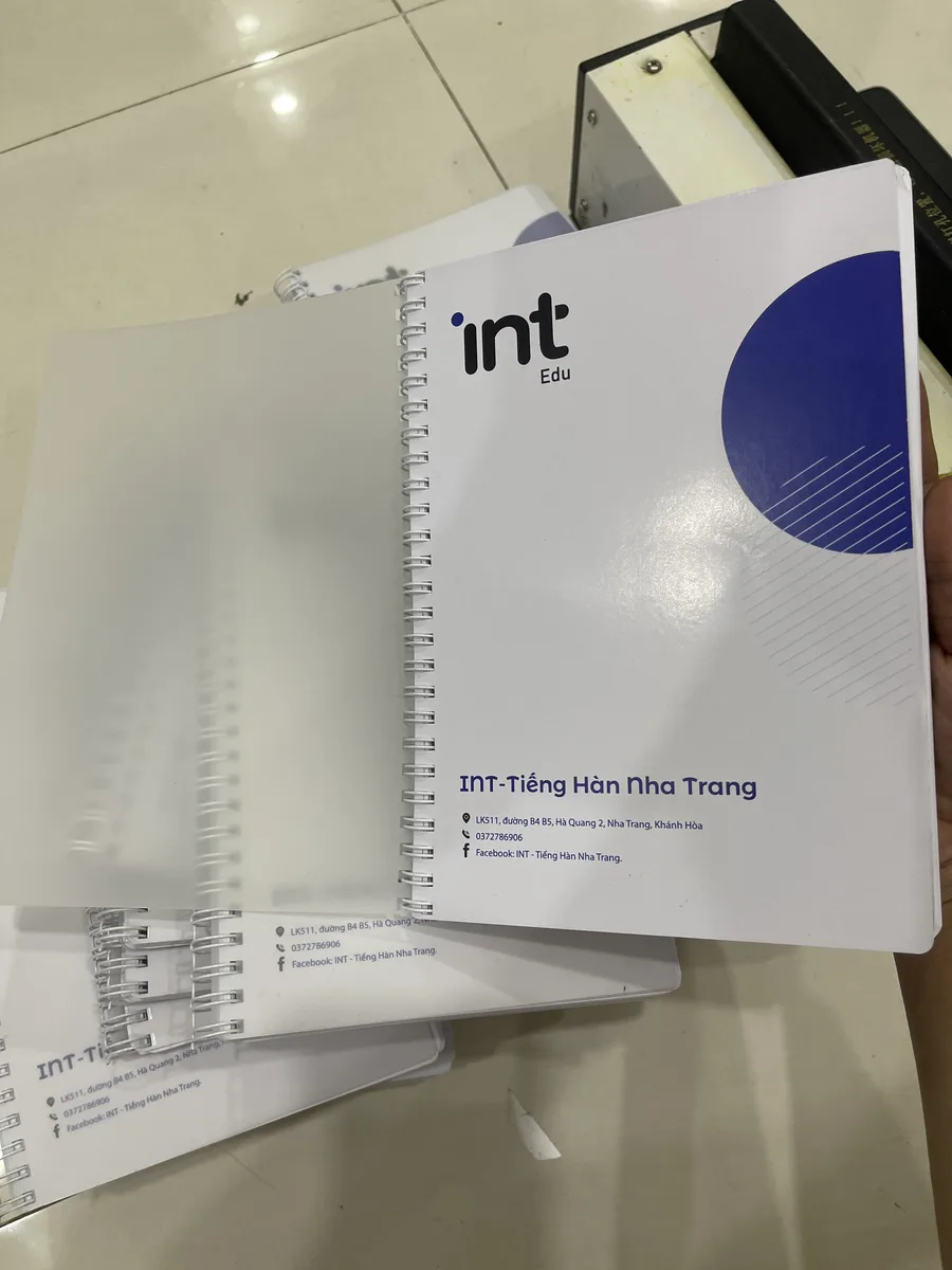 Cuốn sổ tay lò xo mở ra, bìa in logo và thông tin liên hệ của trung tâm ngoại ngữ tại Nha Trang, in tại In Đức Lộc