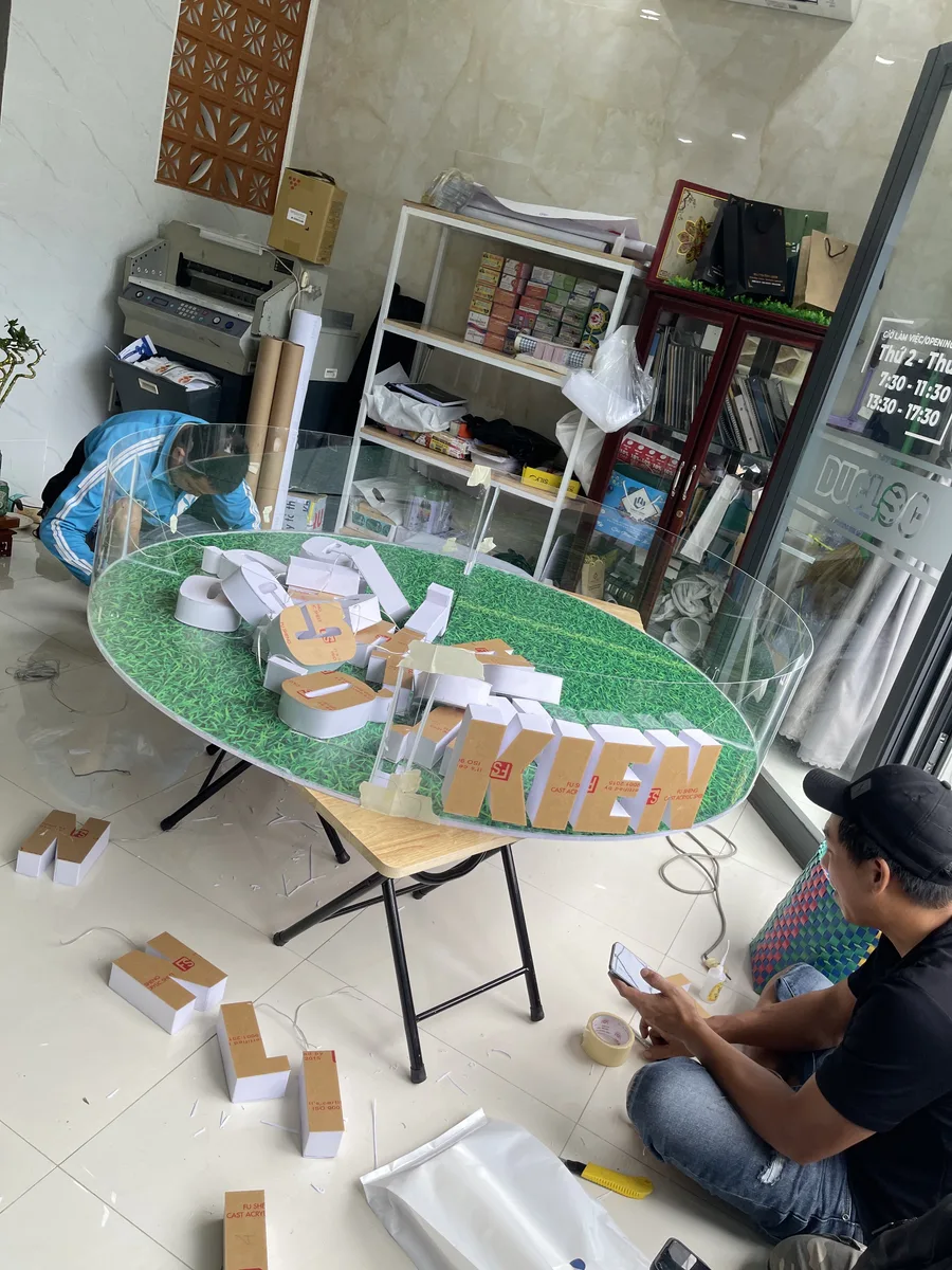 Thợ In Đức Lộc lắp bộ chữ nổi mica trên nền tròn, thấy rõ mặt chữ còn màng bảo vệ và hông chữ trắng